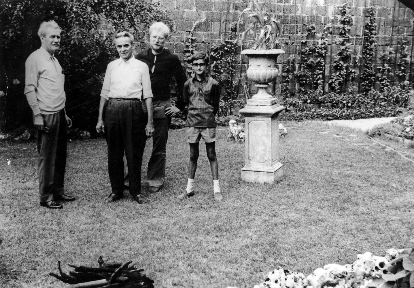 three men and a boy stand on a lawn, beside a large urn